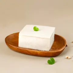 Kostka białego tofu na drewnianej misce, obok nasiona soi. Tofu powstaje z ziaren soi.