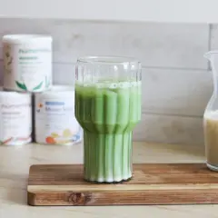 Un verre de matcha latte frais, avec des pots de poudre de matcha et une carafe de lait végétal en arrière-plan.
