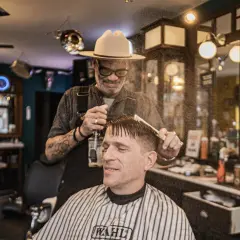 W salonie fryzjerskim "Klasyczna Maszynka" barber w kapeluszu spryskuje wodą włosy klienta przed strzyżeniem.