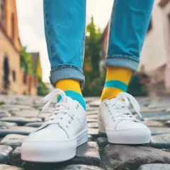 Kolorowe skarpetki do sneakersów dodają charakteru stylizacji. Osoba w jeansach i białych butach idzie po bruku.