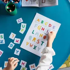 Dziecko układa litery alfabetu na niebieskim stole, obok kolorowanki i kubków z kredkami.