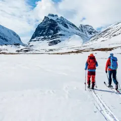 Två skidåkare med ryggsäckar på väg att bestiga berg i snöklädda fjäll under en molnig himmel.