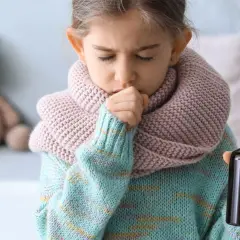 Niña de 3 a 4 años con tos, envuelta en bufanda y suéter, sosteniendo un frasco de medicina.