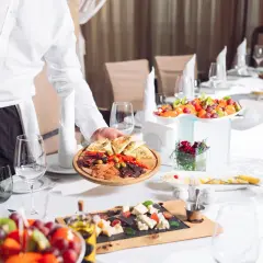 Elegancki kelner serwuje przystawki na stole. Widać owoce, sery i wędliny, co sugeruje wykwintny serwis rosyjski.