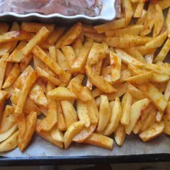 Gotowe do pieczenia frytki w piekarniku, pokrojone w łódeczki i doprawione przyprawami, leżą na blasze wyłożonej papierem.