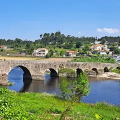 El ponte de Outeiro, un puente de piedra con varios arcos, cruza un río azul. En la orilla opuesta, casas con tejados rojos se asientan en una colina verde.