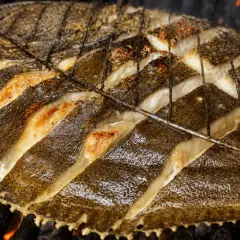 Rodaballo a la plancha, con cortes profundos en su piel oscura, se cocina sobre brasas ardientes, revelando su carne blanca y jugosa.