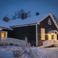 Mörkt trähus med snö på taket och runt om. Varmt ljus lyser från fönster och entré. Skylt med texten "Stilleben" syns.