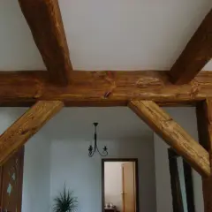 Solidne łączenie belek drewnianych pod kątem prostym, tworzące solidną konstrukcję. W tle widać drzwi i żyrandol.