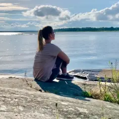 Kvinna sitter på en klippa vid havet, övar mindfulness för att minska stress. Solen glittrar på vattnet.