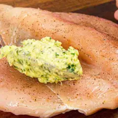 Un cucchiaio di burro alle erbe viene spalmato su un petto di pollo crudo, pronto per essere cucinato.