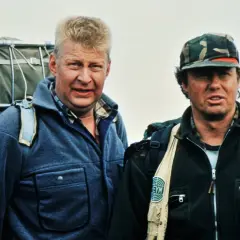 Two men in outdoor gear, one with a large backpack, stand against a pale sky.