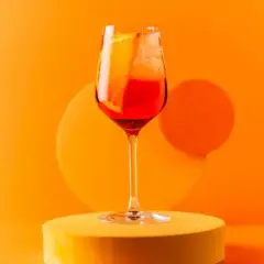 Orzeźwiający spritz campari z plasterkiem pomarańczy i lodem, prezentowany na żółtym podeście na tle pomarańczowego tła.