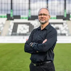 Rafał Smalec, w czarnej koszuli, stoi na tle stadionu piłkarskiego.