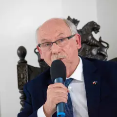 Starszy mężczyzna w okularach i garniturze przemawia do mikrofonu. Na klapie marynarki ma przypinkę z flagami Polski i UE, symbolizującą jego poparcie dla tych, kto głosował za zielonym ładem.