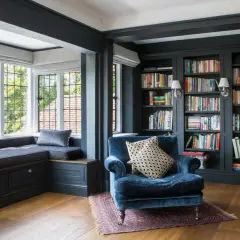 A plush blue armchair sits in a corner cozy reading nook, beside a window seat and floor-to-ceiling bookshelves filled with colorful books.