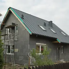 Rozbudowa starego domu w toku. Szary, nowoczesny budynek z metalowym dachem i oknami dachowymi, otoczony rusztowaniami.