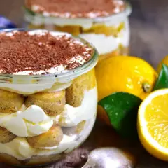 Pyszne tiramisu cytrynowe w słoiczku, przełożone biszkoptami i kremem, posypane czekoladą. Obok świeże cytryny.