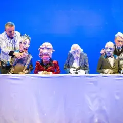 Puppenspieler dirigieren ihre Figuren auf der Bühne im Theater Remscheid. Eine bunte Truppe von Marionetten sitzt hinter einem weißen Tuch.