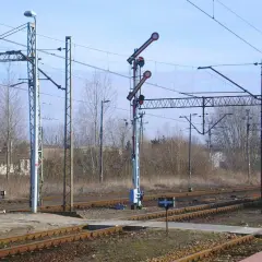 Sygnały na semaforach kolejowych wskazują drogę pociągom. W tle widać komin i drzewa.