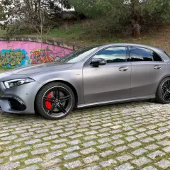 A45 AMG CV gris mate, con pinzas rojas y llantas negras, aparcado sobre adoquines frente a un muro con grafitis.