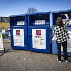 Person lämnar kläder i blå container för textilier återvinning. Skylt visar vad som får lämnas.