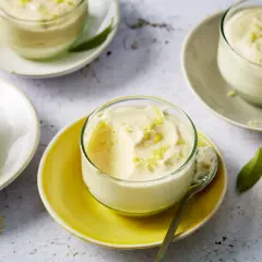 Plusieurs verrines de mousse au citron, garnies de zestes, sur des assiettes claires et jaunes. Un citron coupé est visible à droite.