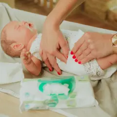 Delikatne dłonie z czerwonymi paznokciami pokazują, jak przewijać noworodka, używając chusteczek nawilżanych.