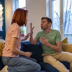 Ein Paar sitzt auf einem Sofa und gestikuliert lebhaft. Sie scheinen sich nach einem Streit wieder zueinander zu finden, die Spannung weicht langsam der Versöhnung.