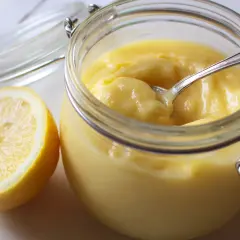 En burk med krämig lemon curd och en halv citron bredvid. En sked vilar i burken, redo att skopa upp den syrliga godsaken.