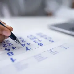 Planowanie urlopu wypoczynkowego. Dłoń z długopisem zaznacza datę na kalendarzu, obok widać laptopa.