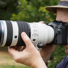 A photographer in a hat uses a large telephoto lens on a Micro Four Thirds camera, perhaps for a detailed micro 4/3 lenses review.