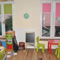 Pokój dziecięcy z zabawkami, stolikiem i krzesełkami. Okna z zielonymi i różowymi roletami, a za nimi widać dachy domów. Trofast inspiracje dla małych artystów.