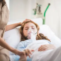 Dziewczynka w szpitalnym łóżku z maską tlenową, walcząca z hipoksją. Opiekunka czule kładzie dłoń na jej głowie.
