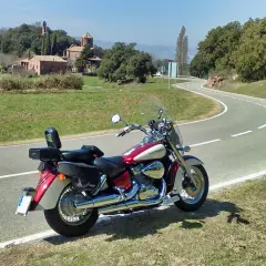 Honda VT 750 C Shadow 2007 ficha técnica. Moto custom roja y crema aparcada en una carretera rural con un paisaje de colinas y edificios antiguos.