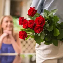 Mężczyzna trzyma bukiet czerwonych róż za plecami, a w tle widać uśmiechniętą kobietę.