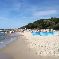 Sianożęty atrakcje: plaża pełna ludzi, falochrony w morzu, zielony las w tle.