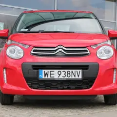 Czerwony Citroen C1 z tablicą rejestracyjną WE 938NV stoi przed budynkiem z dużymi oknami.