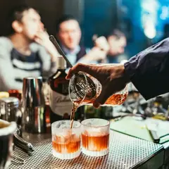 Un barman prépare un cocktail de base, versant un liquide ambré dans deux verres.
