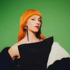 Agnes Carlsson med orange peruk, dramatiska örhängen och en svartvit pälsjacka mot grön bakgrund.