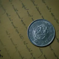 Srebrna moneta bulionowa z wizerunkiem smoka, leżąca na pożółkłym papierze z orientalnym pismem.