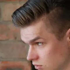 Joven con un corte de pelo moderno, un mid fade en pico, con el cabello peinado hacia arriba y a un lado, frente a una pared de ladrillos.
