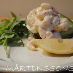 Avokadohalva fylld med räkor i en krämig sås, liknande guacamole crème fraiche, med en citronskiva och gröna blad.