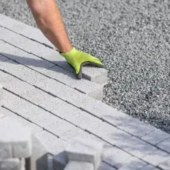 Ręka w zielonej rękawicy układa kostkę brukową na podsypce cementowo piaskowej.