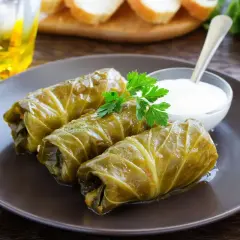 Tre involtini di cavolo cappuccio, guarniti con prezzemolo, serviti con una ciotolina di salsa bianca e un bicchiere di bevanda dorata.