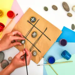Mani che creano un gioco del tris fai da te dipingendo simboli su sassi. Materiali colorati e pennelli completano la scena.