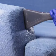 Blaue Polster reinigen mit Rasierschaum. Ein Handschuh hält ein Reinigungsgerät, das Schaum auf den Stoff sprüht.