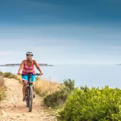 Woman enjoys cycling tours in Slovenia, biking along a coastal path with the sea and a distant lighthouse.
