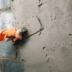 Mano con guante naranja aplica cemento a una fachada enfoscada con llana.