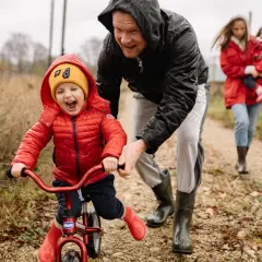 Radosne dziecko na rowerku biegowym, kiedy dziecko stoi bez podparcia, z tatą asekurującym. W tle mama z drugim dzieckiem.